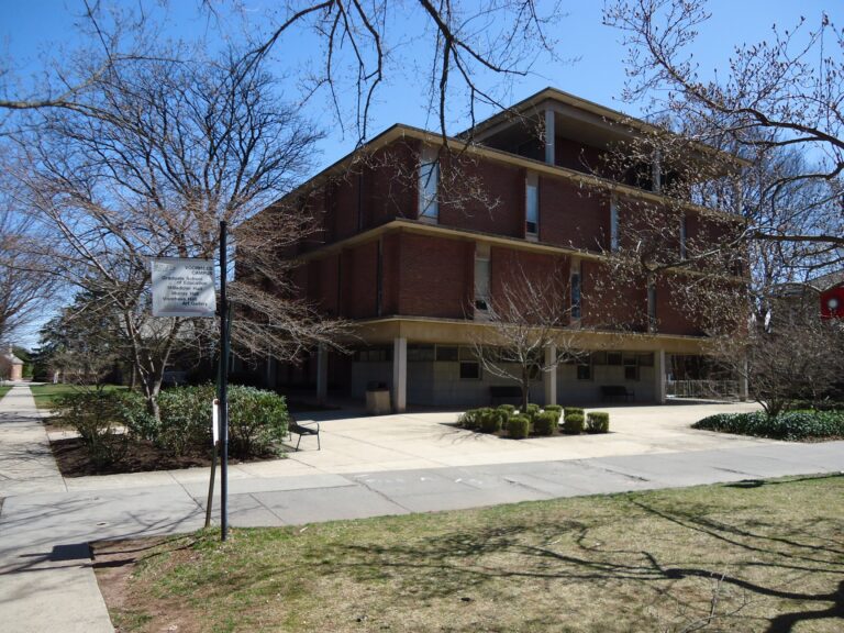 Edificio universitario de ladrillo con ventanas altas y varios estudiantes cruzando la entrada