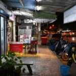Fachada de una pequeña tienda de barrio con el escaparate iluminado y cartel de horario