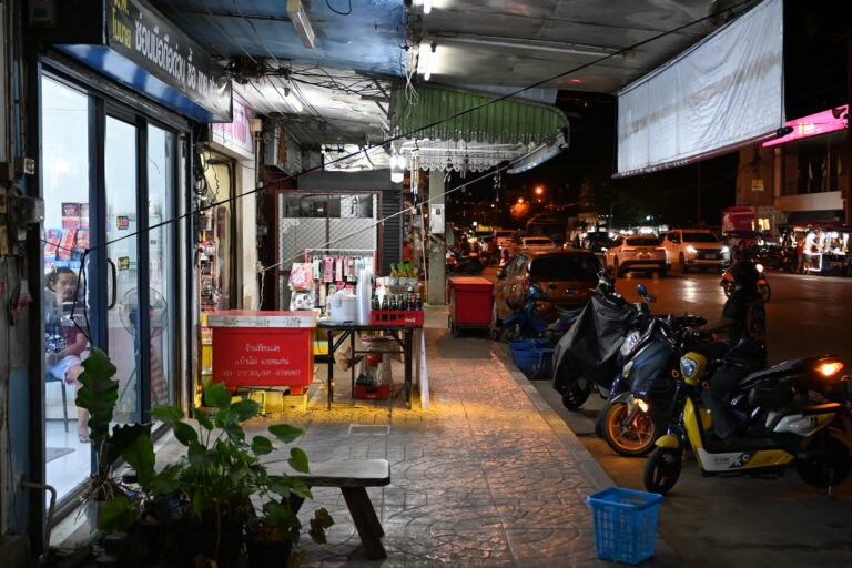 Fachada de una pequeña tienda de barrio con el escaparate iluminado y cartel de horario
